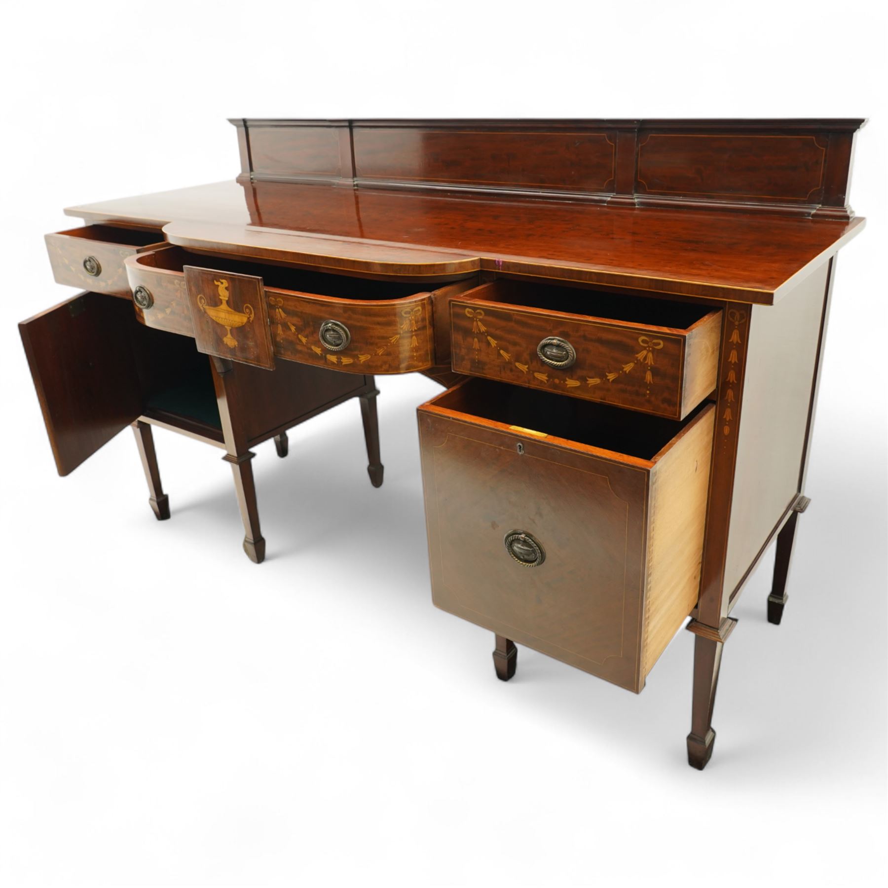 Edwardian Sheraton Revival inlaid plum pudding mahogany serpentine sideboard, rectangular top with raised gallery above bowed frieze drawer finely inlaid with central neoclassical urn motif, flanked by two drawers with swags and ribbon bows, above a cellarette drawer and cupboard, on square tapered supports with spade feet 