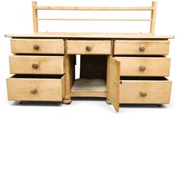 Victorian stripped pine dresser base with plate rack, the four-tier plate rack with supporting rails and hooks, the inverted-breakfront base fitted with seven assorted graduating drawers surrounding a recessed cupboard, on compressed bun feet with castors