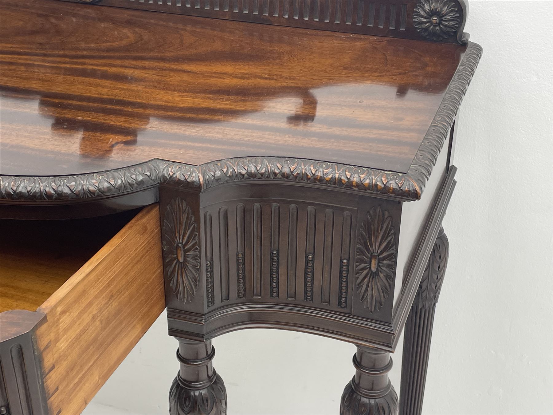 Early 20th century Hepplewhite style serving table, raised fleur-de-lis feather back with fluted detail and flower head mounts, the shaped top with carved foliate edge over single central drawer with applied urn motif, turned and reeded acanthus supports with recessed castors