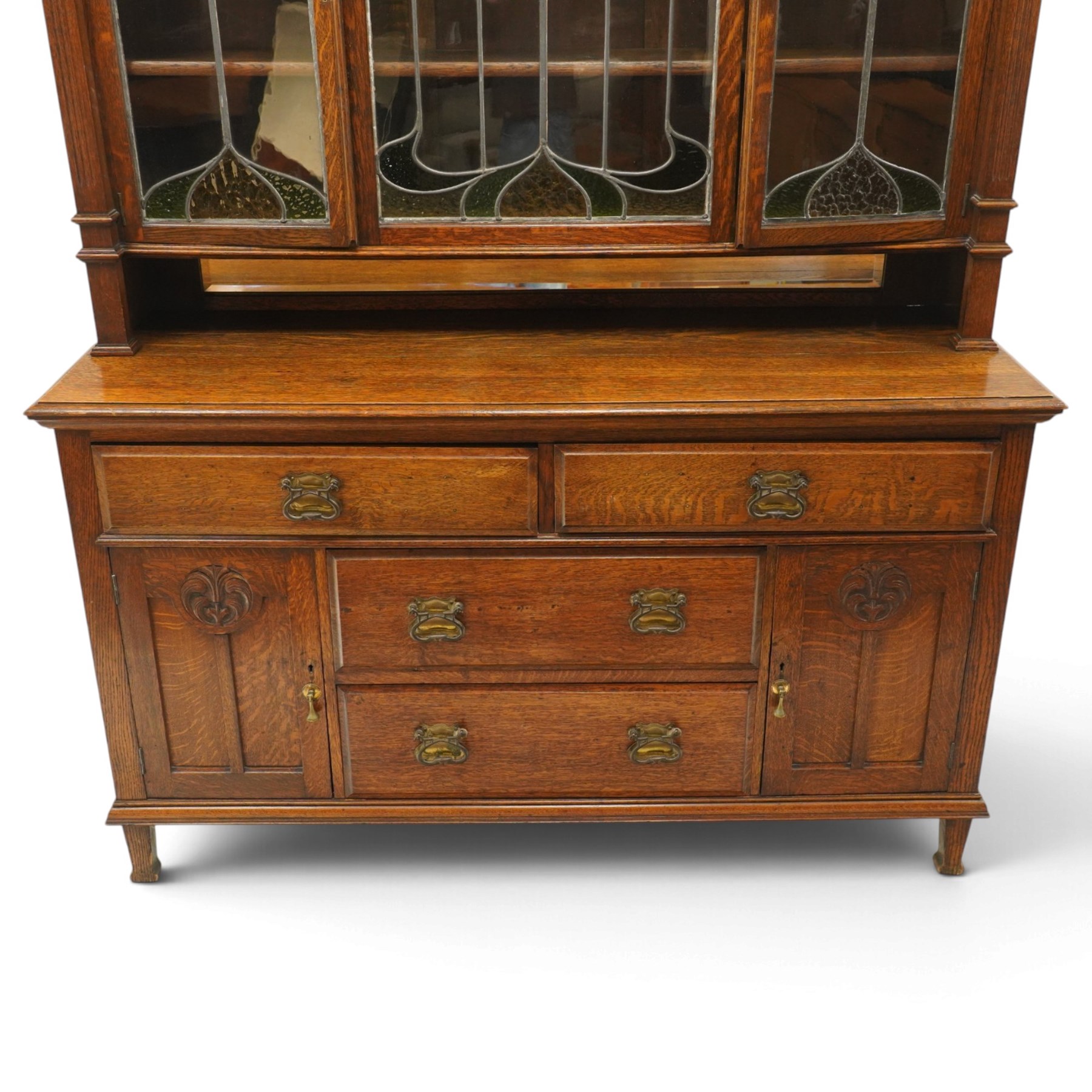 Arts and Crafts oak dresser, projecting moulded cornice above three glazed panels with coloured leaded glass floral panels, flanked by two glazed doors enclosing two adjustable shelves, open mirrored recess below with mirrored back, the base fitted with two short drawers over two central drawers flanked by carved panelled cupboard doors, ornate brass handles, raised on tapering square legs