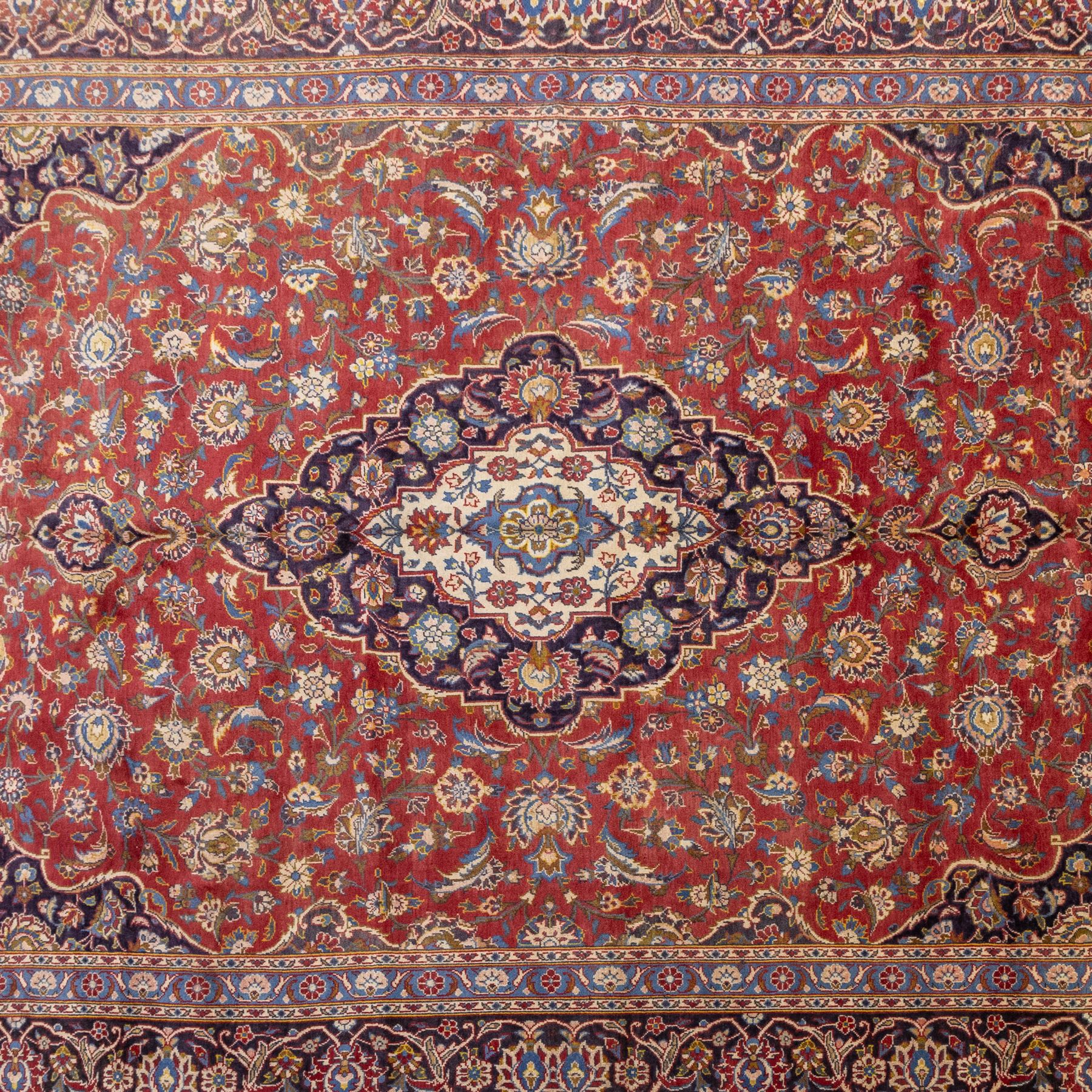 Central Persian Kashan hand-knotted carpet, madder red field with a central cusped medallion of ivory and deep indigo ground, enclosing a stylized floral rosette, framed by scrolling arabesques, field richly ornamented with an intricate network of flowering vinery, palmettes, and delicate floral sprays, spandrels in deep indigo, principal border of navy ground decorated with a continuous palmette and vine-scroll, flanked by multiple guard stripes, with cotton fringe ends 