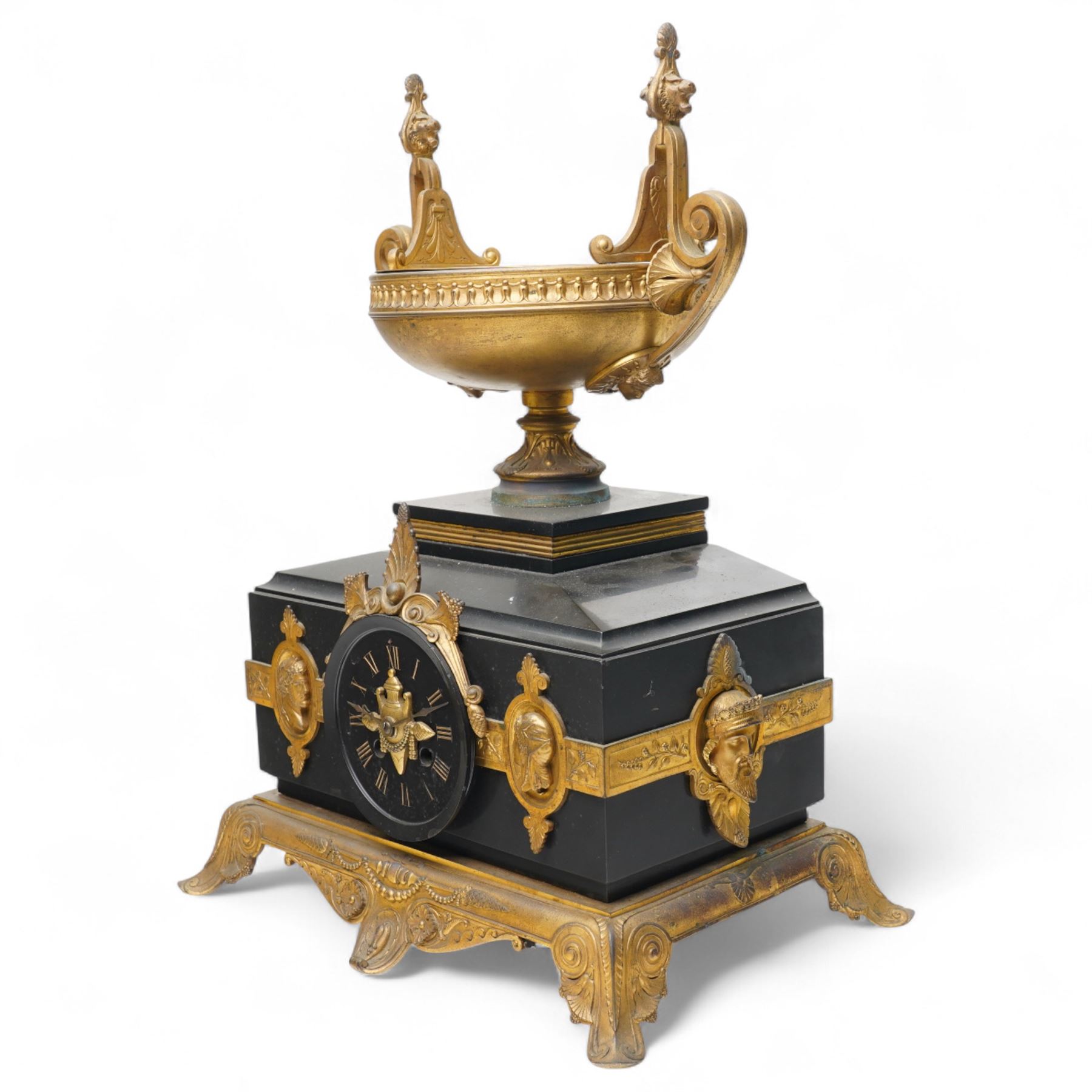 French- late 19th century 8-day mantle clock in an imposing Belgium slate case with gilt metal ornamentation. Stepped rectangular case with an angled top and square plinth surmounted by an oval shaped gilt urn with scroll handles, case with continuous strapwork and oval medallions displaying portraits from classical antiquity, raised on a gilt metal base with splayed feet, conforming unenclosed slate dial with gold incised Roman numerals and brass Fleur de Lis hands, Parisian twin train rack striking movement with pendulum regulation, striking the hours and half hours on a bell. With pendulum.