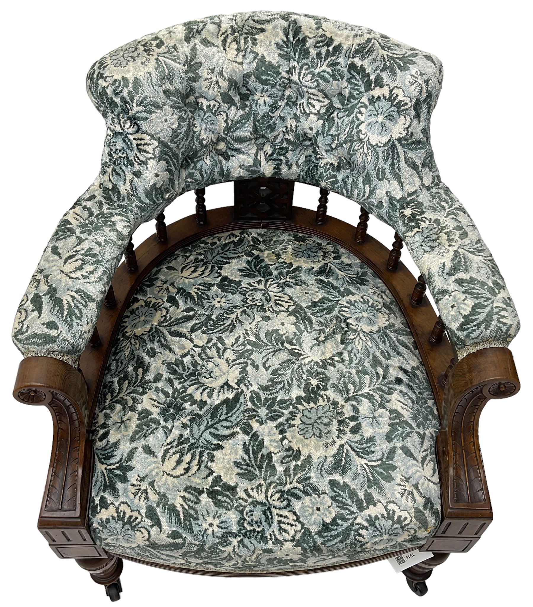 Victorian walnut framed tub-shaped armchair, the shaped rolled back supported by a spindle turned balustrade, upholstered in buttoned floral fabric, scrolled arms supports carved with acanthus leaves and flower heads, on collar turned feet with brass and ceramic castors
