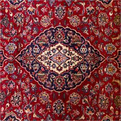 Central Persian Kashan hand-knotted rug, crimson red field with central ivory and indigo floral medallion surrounded by an all-over arrangement of finely drawn palmettes, flowering vines, and rosettes, with corresponding navy blue spandrels enclosed by a floral main border with multiple guard stripes, with cotton fringe ends    