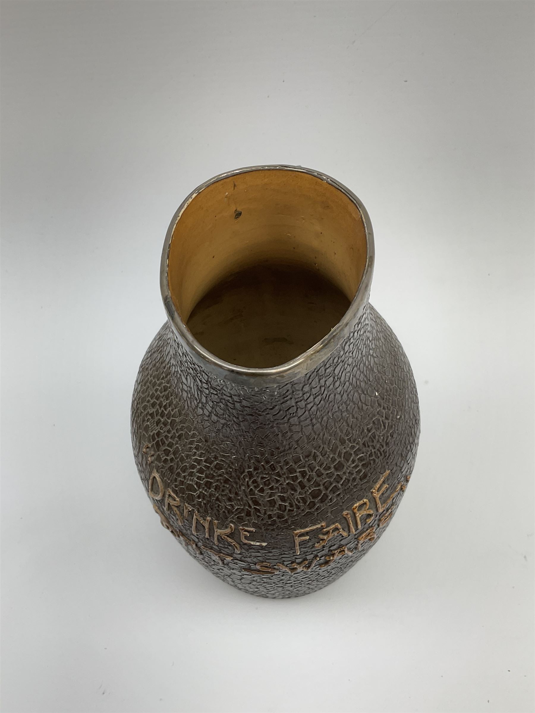 A Doulton Lambeth stoneware jug, with silver mounted collar, hallmarks rubbed, maker's mark probably Cornelius Desormeaux Saunders & James Francis Hollings (Frank) Shepherd, the leather effect body titled 'Drinke Faire Don't Sware', with impressed marks beneath including 'Manufactured for Sidney W Allen 39 White Rock Hastings', H18.5cm. 