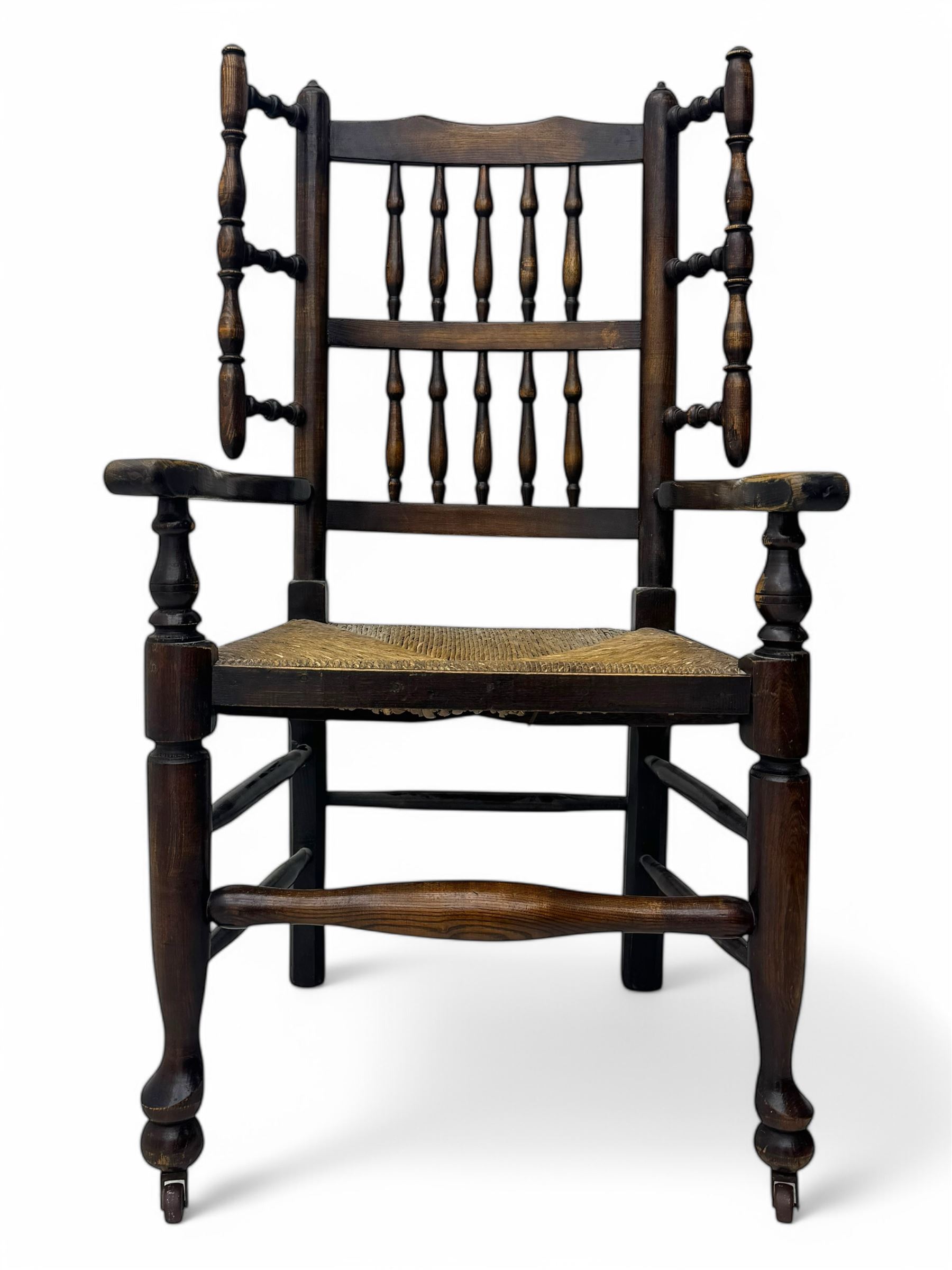 Early 19th century ash armchair, the back with winged spindles extending from a shaped top rail, above a splat of two rows of vertical spindles, with curved arms and a rush seat, raised on turned supports united by double stretchers terminating in castors