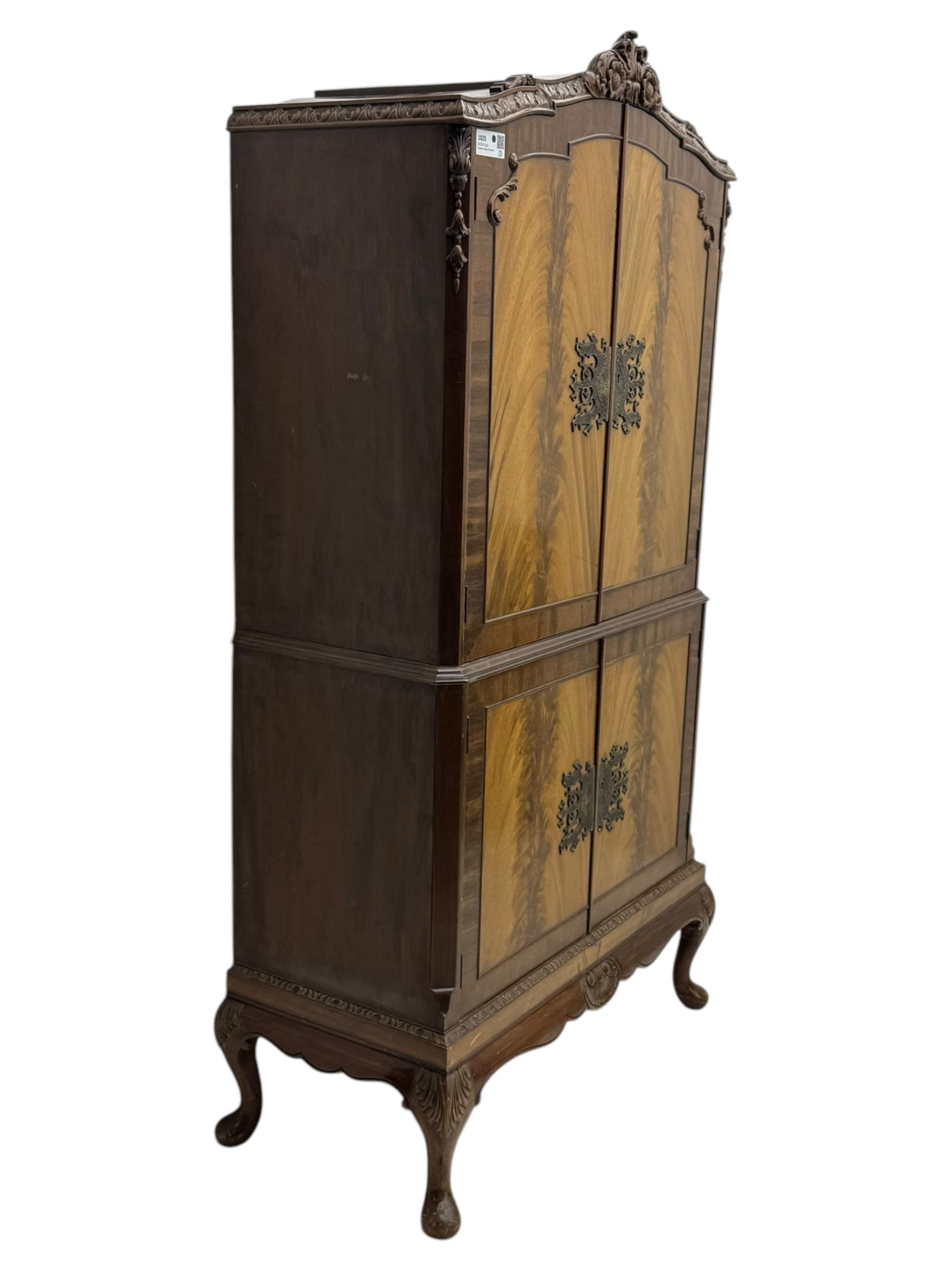 Early 20th century walnut veneered cocktail cabinet, shaped and carved foliate pediment above a pair of arched panelled doors with figured veneers and ornate cast escutcheons, interior is mirrored on the base and back, slide out mirrored shelf, flanked by foliate carved uprights, over a conforming lower cabinet, all raised on shaped apron and scroll carved cabriole feet