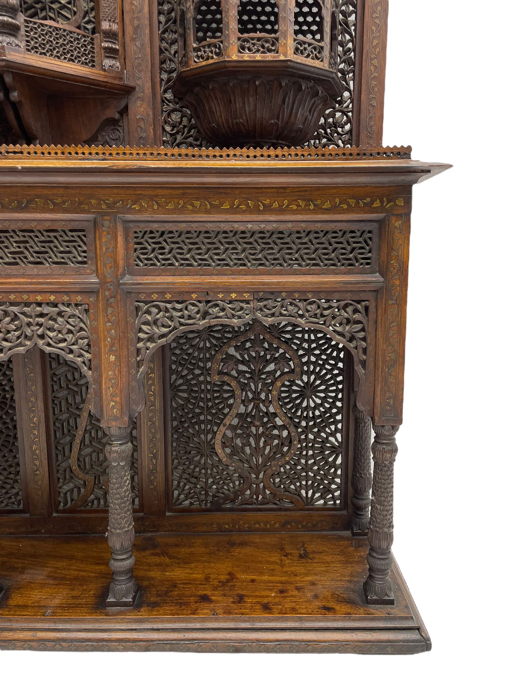 Late 19th century Indian Burmese carved and pierced hardwood and brass inlaid wall shelf, the shaped back profusely decorated with trailing and interlaced foliage, with projecting architectural structures, the central balcony with canopy top and five open windows with arched apertures on columns, two flanking five-sided balconies, the shelf supported by six foliate carved columns with pierced cusped pointed arches 