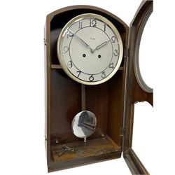 A mid 20th century German wall clock in a veneered mahogany case with a German eight day 'Kienzle' movement sounding the hours and half hours on twin gong rods, 9' circular dial with chrome Arabic numerals and pierced baton hands,  full length door with glazed dial and pendulum apertures, chrome pendulum bob. 
 


