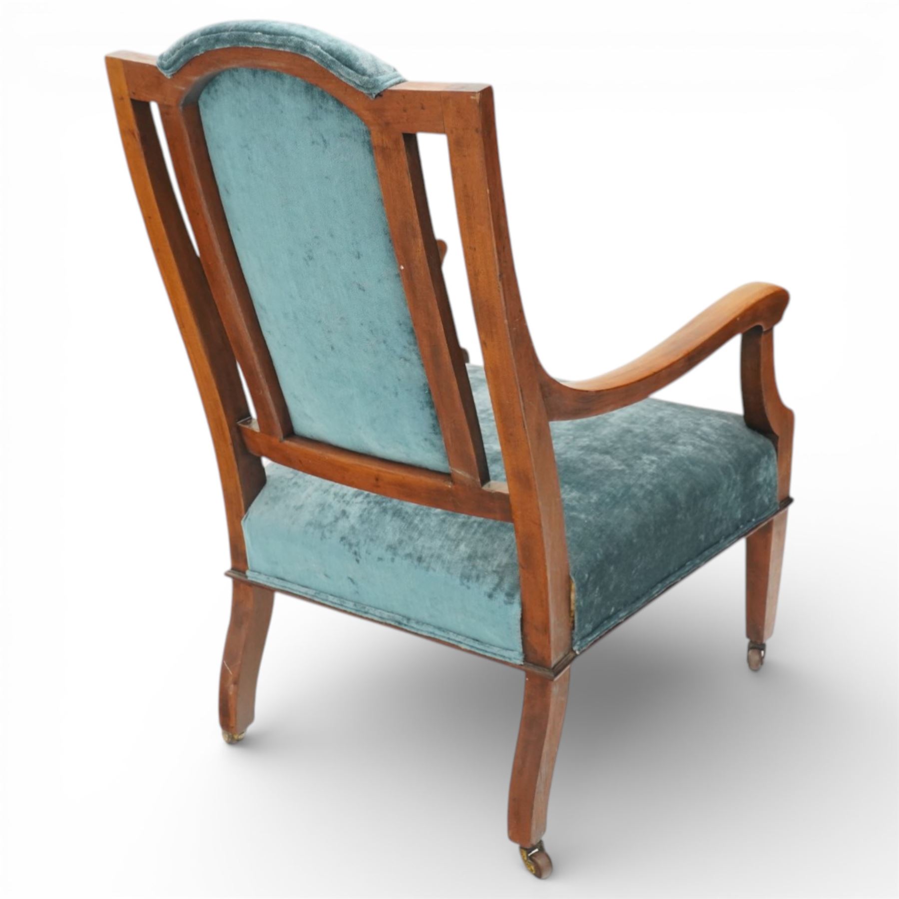 Arts & Crafts mahogany framed armchair, shaped rectangular back with open uprights, padded back, seat and arm supports upholstered in blue velvet, downswept arms on square tapering front supports with brass caps and castors, the rear supports slightly splayed