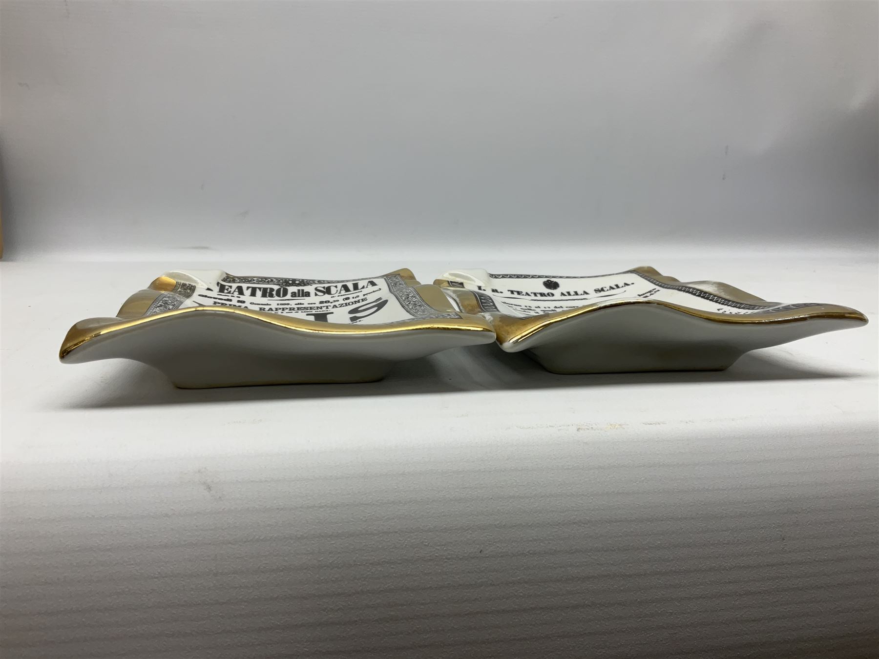 Two Fornasetti rectangular operatic posters ashtrays 'Norma' and 'Iris' decorated with black and white text with a gilt border edge, with printed mark beneath, H16cm