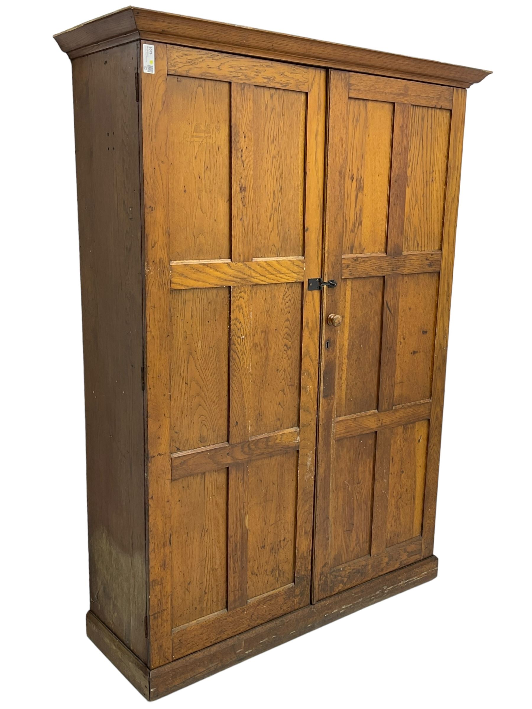 The North of England School Furnishing Company Limited Darlington - early 20th century oak school cupboard, projecting moulded cornice over two panelled doors, fitted with shelves, on chamfered plinth base 