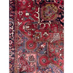 Northwestern Persian Heriz hand-knotted carpet, madder red field with a bold stepped medallion in indigo, ivory, and ivory, surrounded by angular floral motifs and stylized leaves, and the spandrels set within a dark border of rosettes and angular vine motifs, 335cmx252cm