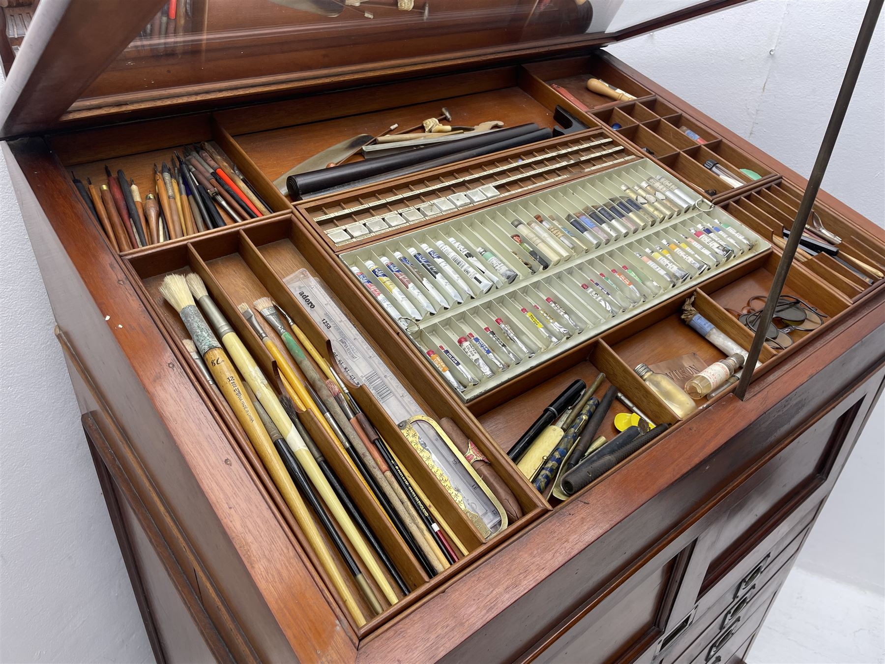 Edwardian mahogany artist's cabinet once belonging to the British artist Eli Marsden Wilson (1877-1965), raised panelled back mounted by carved brackets, sloped and hinged glazed lid enclosing paint holders and compartments, up-and-over panelled door revealing three slides, fitted with two banks of five graduating drawers with recessed brass handles, on skirted base.

Together with a quantity of artists' equipment and tools including paints, paintbrushes, charcoal, chalks, etc. and various prints relating to the life of the artist, including Quaker anti-war posters, Victorian Stevengraphs, and various chromolithographs 

Provenance: Removed from the studio of Eli Marsden Wilson. The cabinet comes to us for sale through family descent. The vendor is descendant of Marsden. Many further etchings removed from the cabinet are being sold in the Spring Art Sale, Friday 15th March 2024, Lots 265-271.

Notes: born in Ossett, Eli Marsden Wilson was the only son of Alfred Wilson, a foreman beamer, and Emma Marsden, and had five sisters. After studying at Wakefield College of Art, he moved to the Royal College of Art in London, where he became a pupil of Sir Frank Short. He had his first exhibit at the Royal Academy in 1905, the same year he married fellow artist Hilda Mary Pemberton.

A Quaker, vegetarian, and pacifist, he was a conscientious objector in World War I and as such was imprisoned in 1917 for two years. In September 1922 he was commissioned by Princess Marie Louise to produce miniature etchings for Queen Mary's Dolls' House at Windsor Castle; two such etchings are being sold in the Spring Art Sale, Friday 15th March 2024, Lot 265.