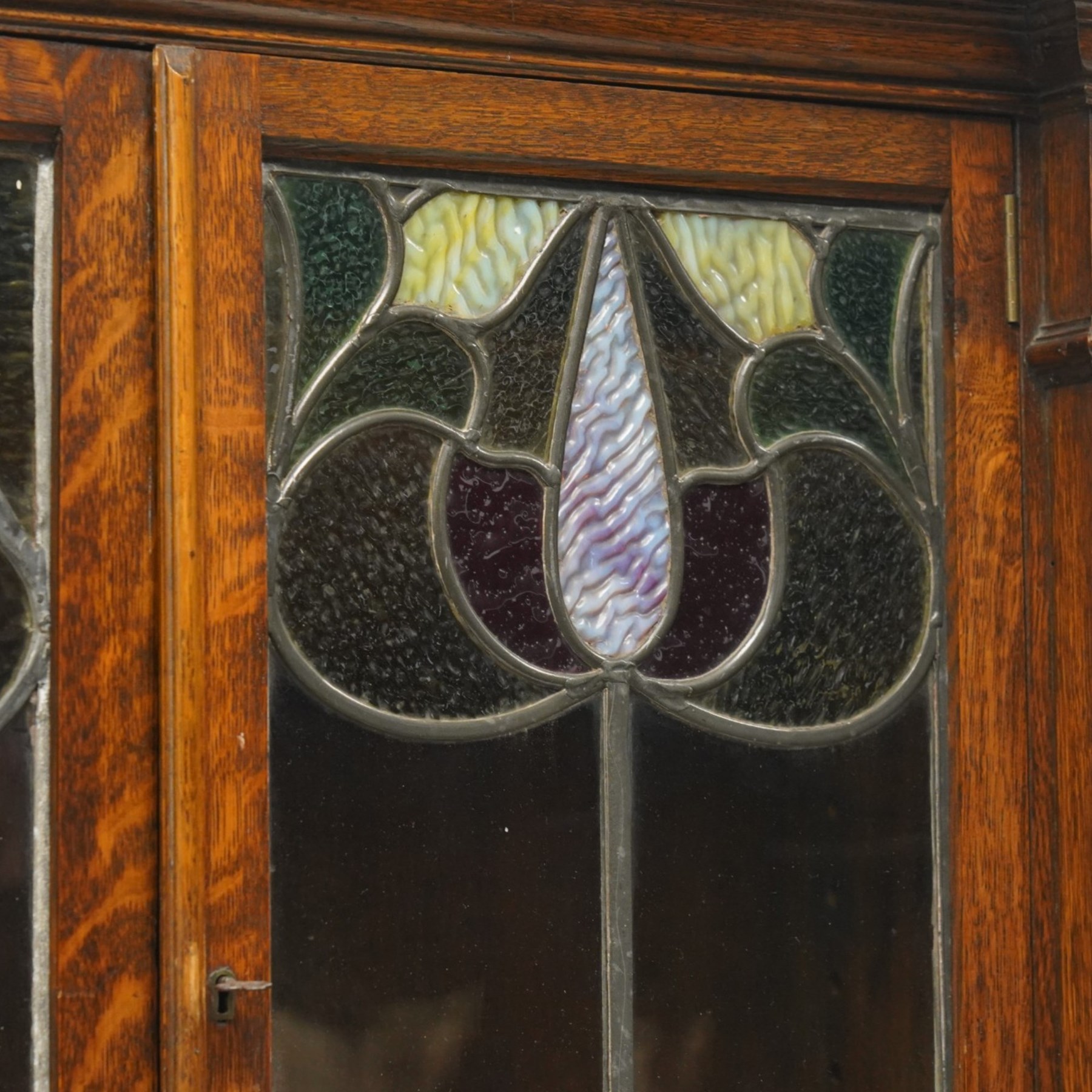 Arts and Crafts oak dresser, projecting moulded cornice above three glazed panels with coloured leaded glass floral panels, flanked by two glazed doors enclosing two adjustable shelves, open mirrored recess below with mirrored back, the base fitted with two short drawers over two central drawers flanked by carved panelled cupboard doors, ornate brass handles, raised on tapering square legs