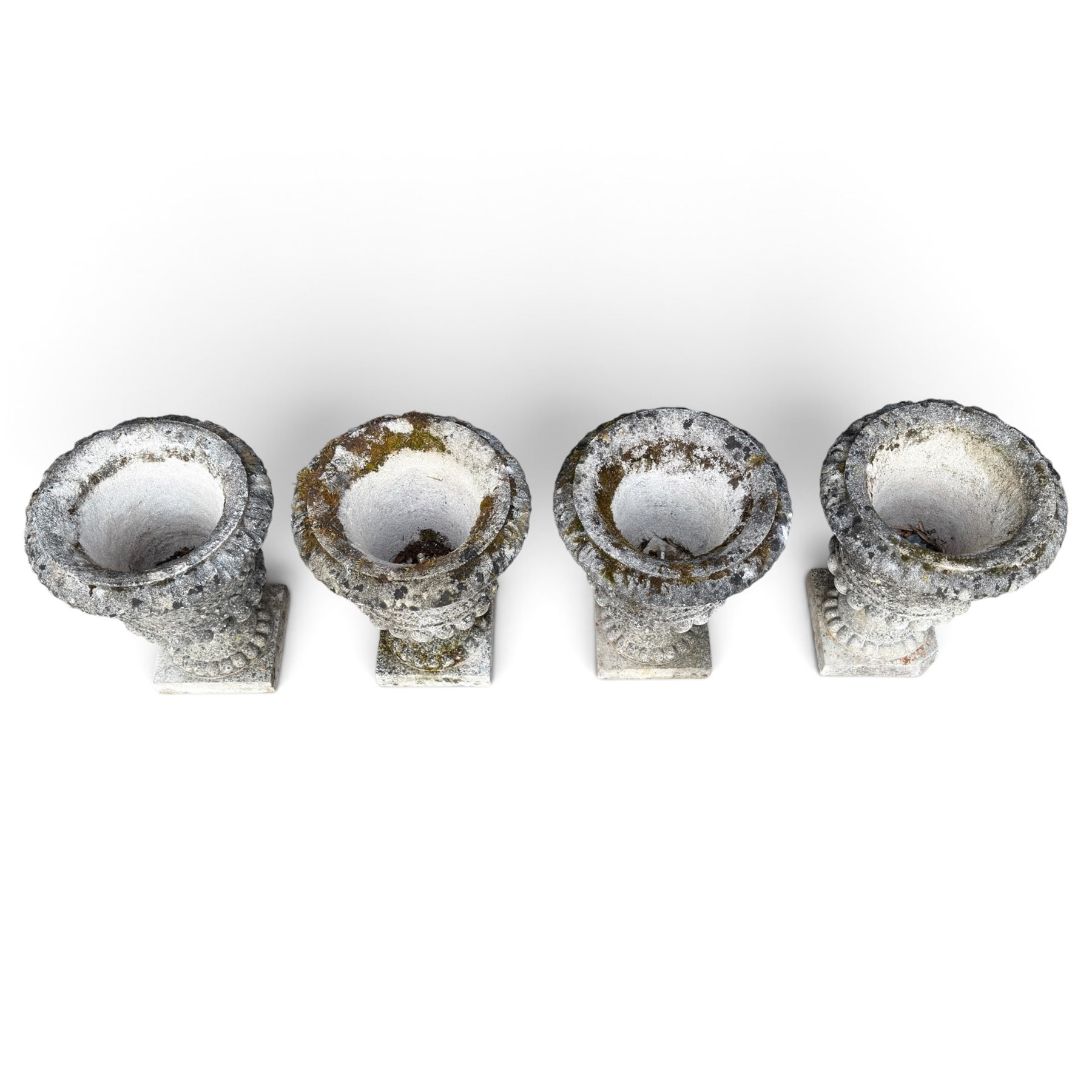 Set of four weathered composite stone urns of campana form, the everted leaf-moulded rolled rim over a body decorated with scrolling fruiting grapevines in relief, over a gadrooned underbelly and waisted socle, lobed collar on square base