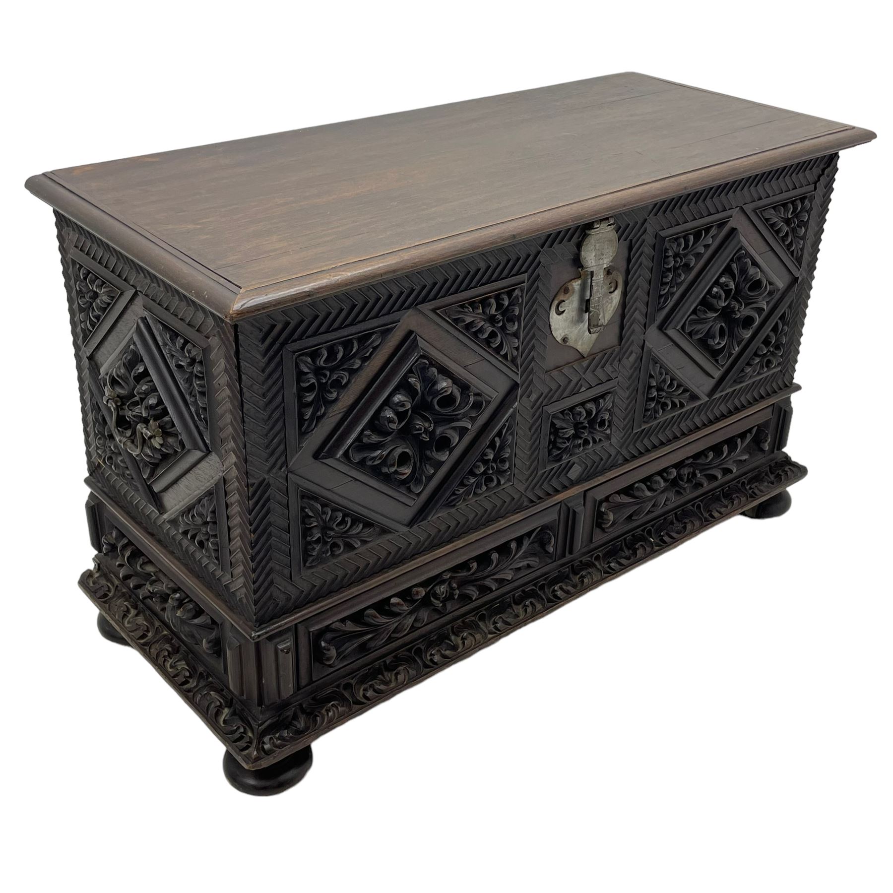 Early 20th century carved hardwood kist or mule chest of small proportions, moulded rectangular hinged lid over foliage carved geometric lozenge panels, the frame carved with chevron banding, fitted with two drawers, the drawer fronts carved with extending leaf motifs over a foliage carved lower moulding, on turned feet
