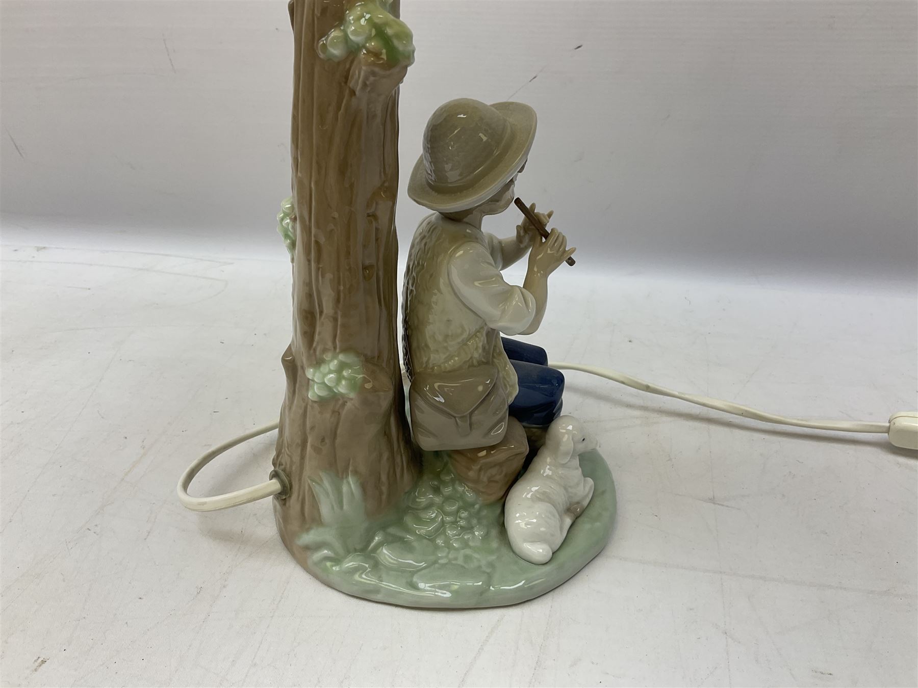Nao table lamp modelled as a boy playing the flute upon logs with two recumbent sheep at his feet, the stem modelled as a tree trunk, with fabric shade, with printed mark beneath, H46cm